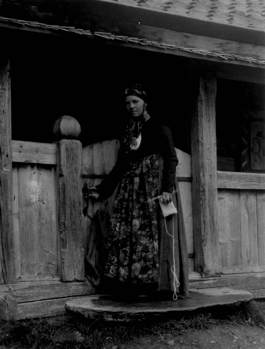 Kvinnedrakt fra Rust, Eggedal. Over 100 år gammel. Kvinne med strikketøy foan svalgang.
