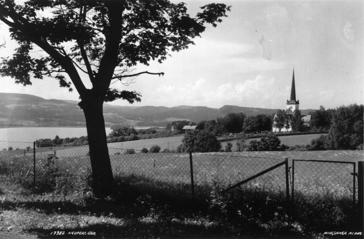 Ringsaker kirke, eksteriør, juni 1935. Jordbruksbygd med gårdsanlegg. Mjøsa, skog og åser i bakgrunnen.