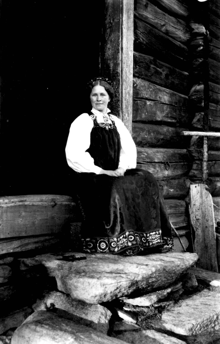 Kvinnedrakt, Ål eller Hol, Hallingdal. Kvinne sittende i åpningen til et tømmerhus.