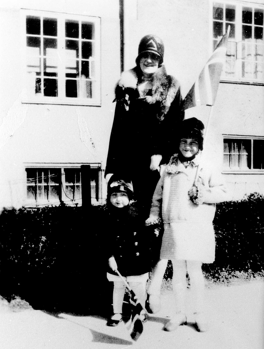Mor, datter og nabojente foran familiens hjem i Jacob Aalls gate. 60, Oslo 17. mai 1930. Mor med korsrev rundt halsen, datter med rød pjekkert og svart silkehatt med blomster på bremmen.
Fra Jorunn Fossbergs familiealbum.