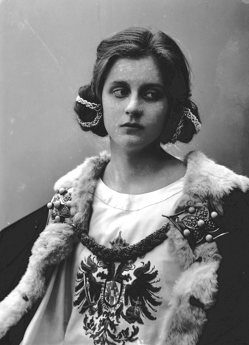 Studioportrett av kvinne i kostyme og oppsatt hår - overdel med riksvåpen og perler i håret.