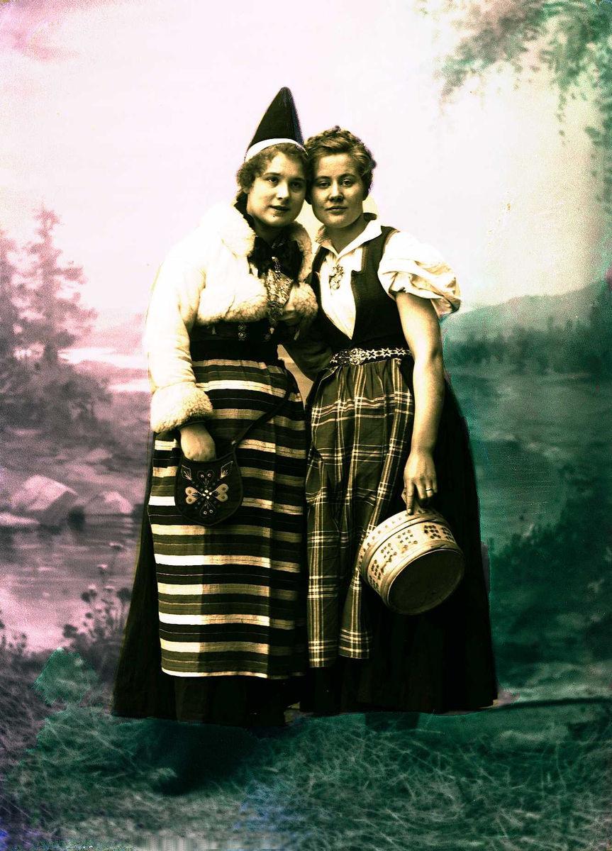 To kvinner i drakt holder rundt hverandre. En holder en ambar i hånden. Malt bakgrunn med landskap, høy på gulvet foran teppet.
