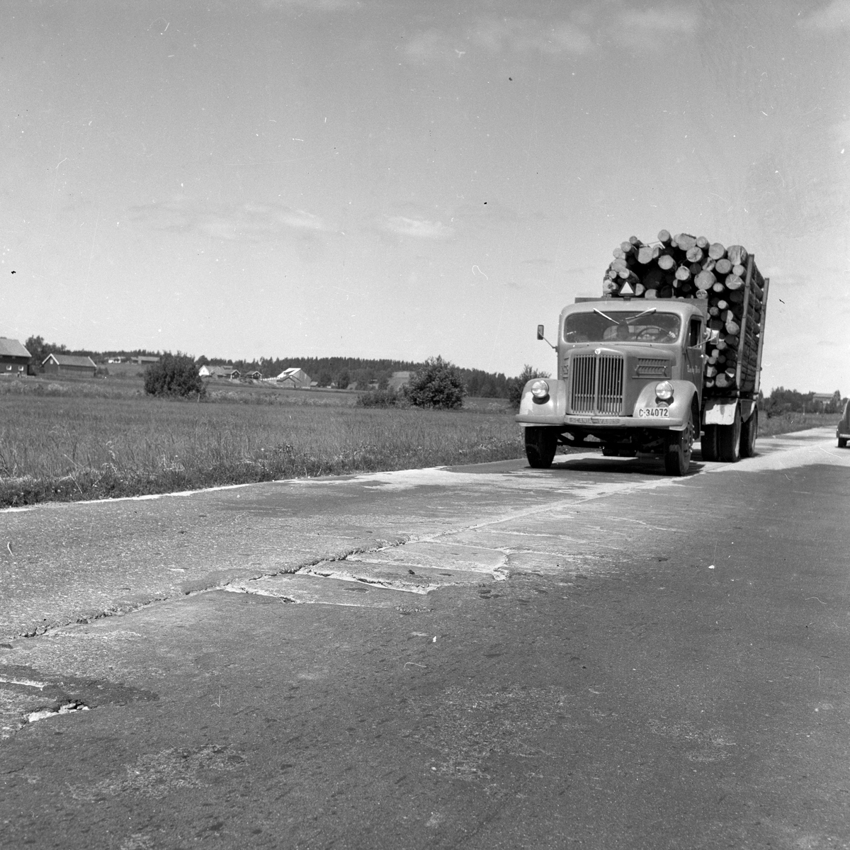 Langs Riksvei 1 kjører en lastebil med tømmerlast i et åpent landskap med bondegårder. Fotografert 7. juli 1958.
