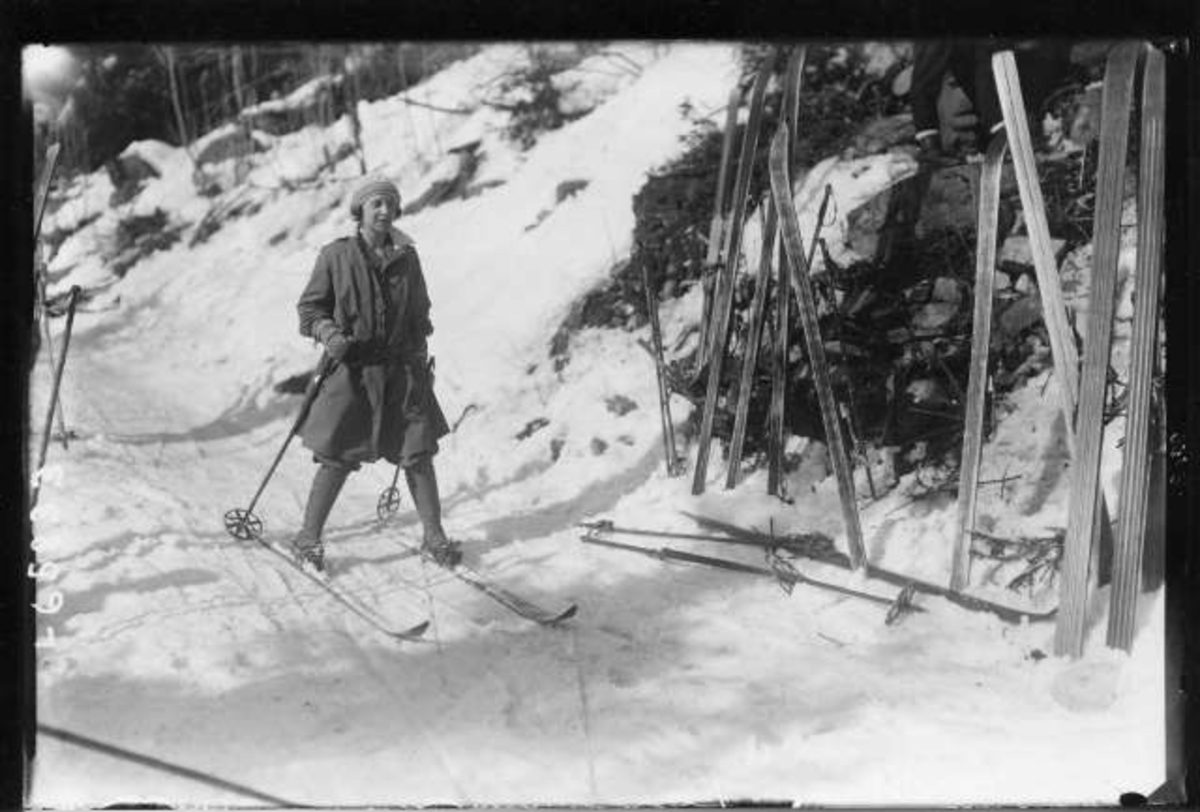 Kvinne på ski ankommer rasteplass med flere par ski stående i snøen.