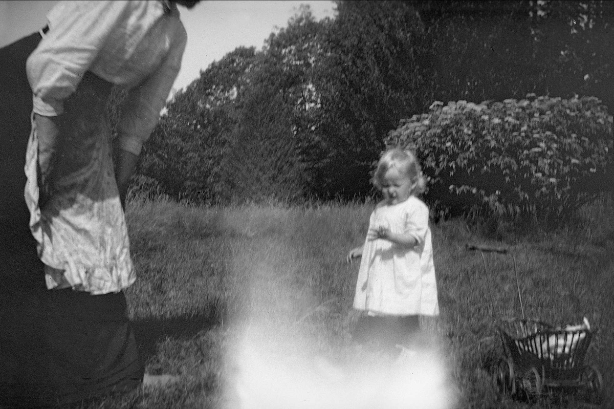 En jente står foran en trillevogn og viser noe til en kvinne. Robsahm og Lund.