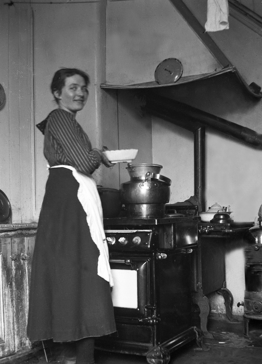 Hushjelpen i kjøkkenet på Elverhøy, Trondheim. Elektrisk komfyr med varmtvannskjele oppå. Til høyre kobberkjele for varmtvann til bad, fyrt med ved og koks. Fotografert 1919.