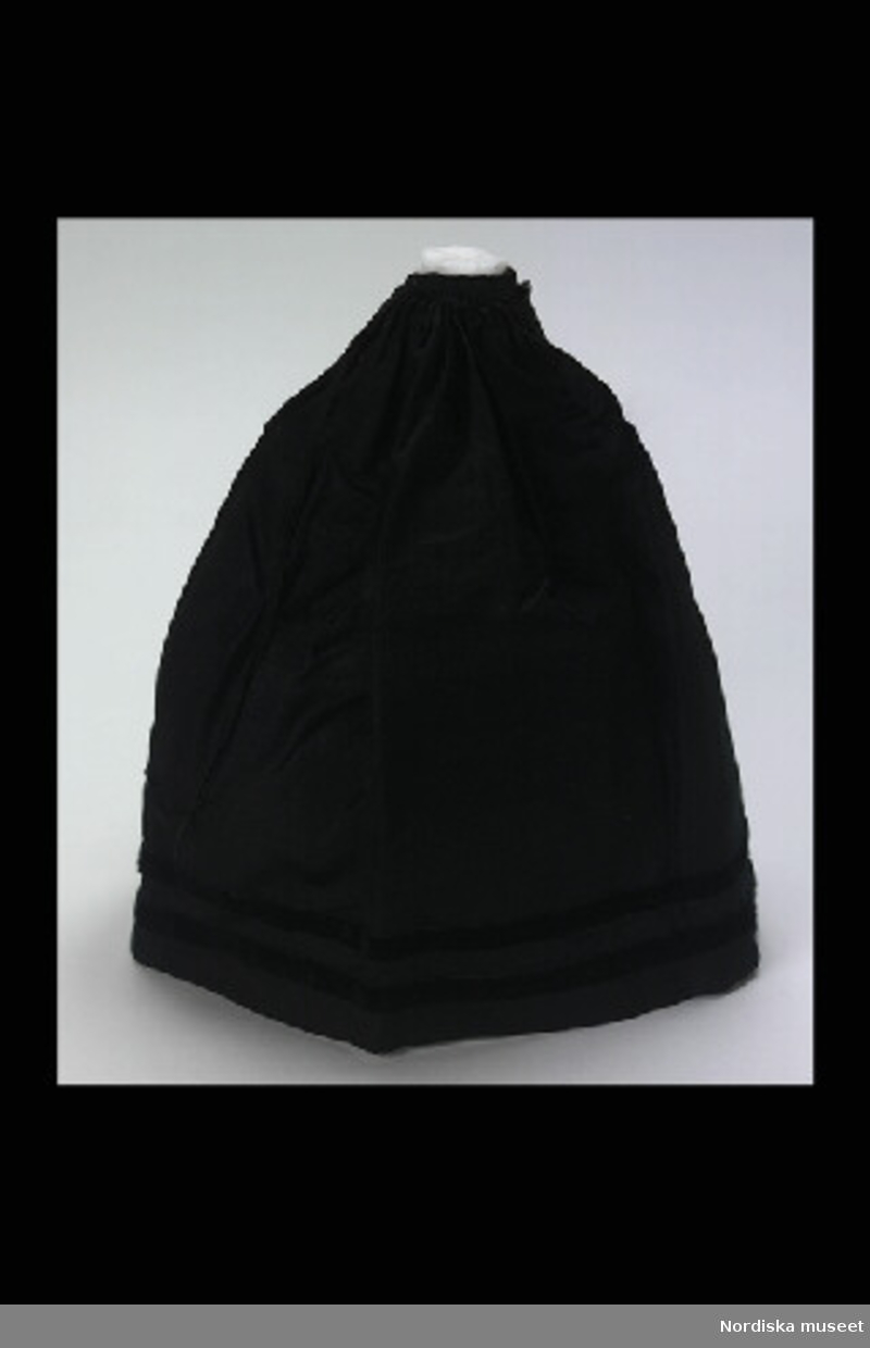 Inventering Sesam 1996-1999:
L 23 (cm)
Förkläde, dockförkläde, av svart sidensatin, dekorerat med svarta sammetsband, veckad linning med två svarta glasknappar och bomullsband.
Anna Womack 1996