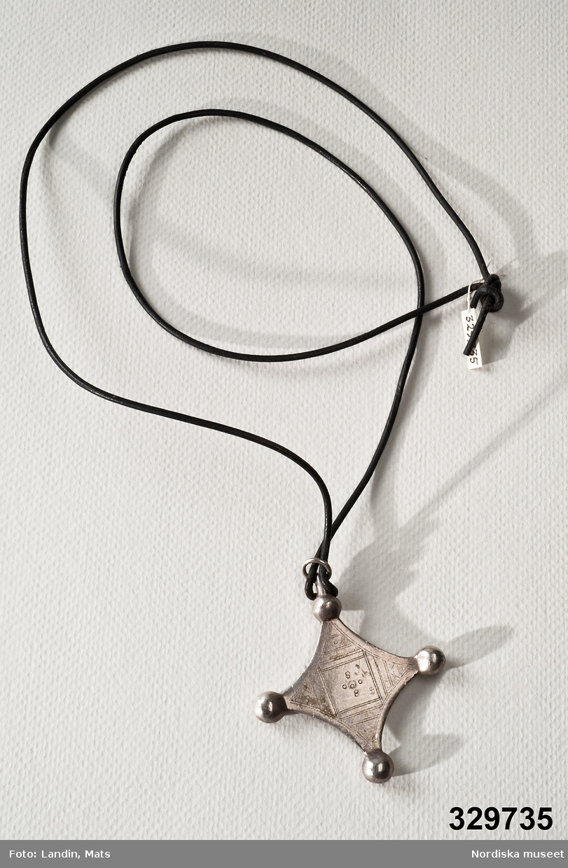 Halsband av smal svart läderrem med hänge i silverfärgad metall, korsformig med kulor i ändarna. Stämpel med texten "Scooter Paris" på baksidan.
/Zingoalla Rosenqvist 2009-02-05