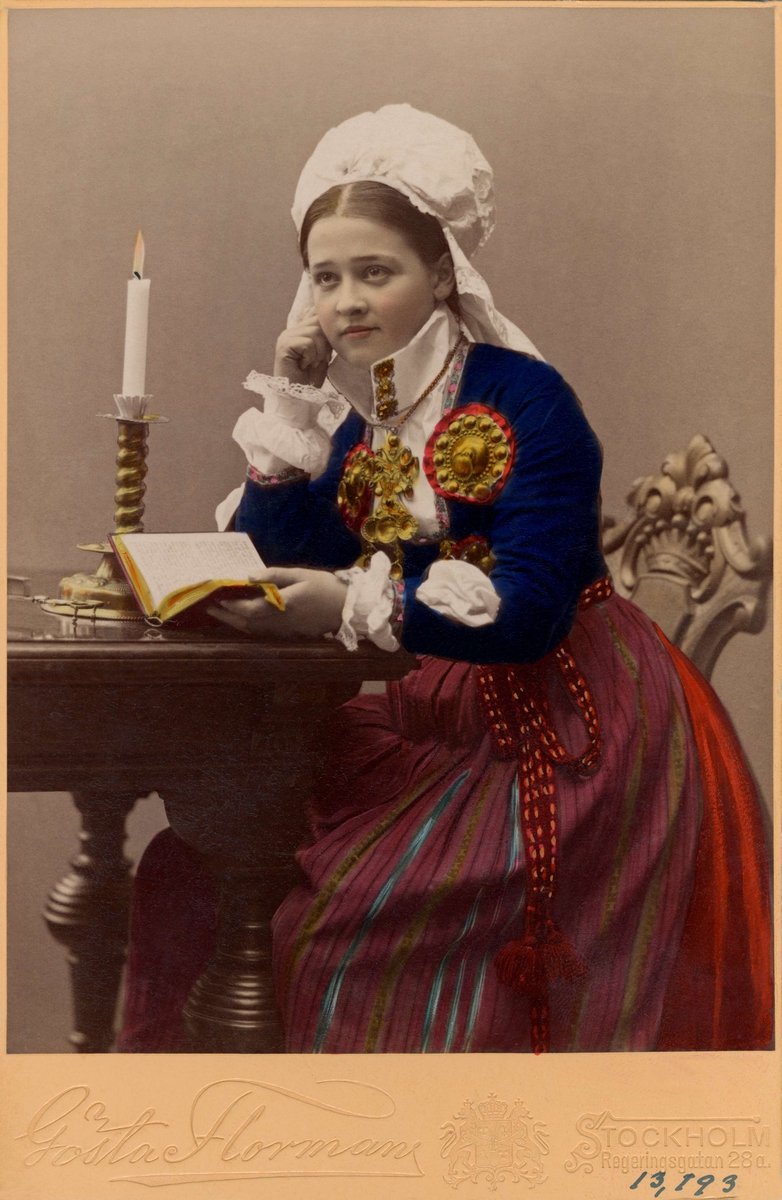En ung flicka i dräkt från Herrestad eller Järrestad, Skåne, sitter vid ett bord med en bok.