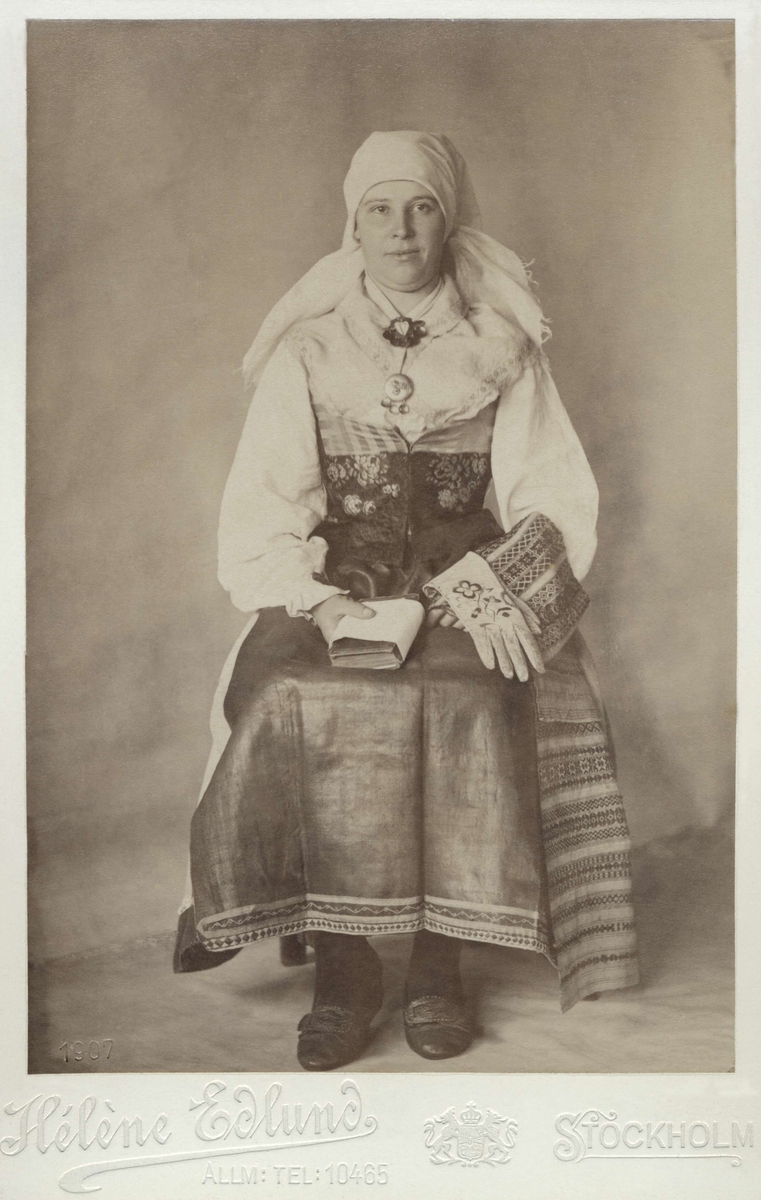 Kyrk och gillesdräkt från Norra Öland för gift kvinna. Porträtt av sittande kvinna med psalmbok och broderade handskar i knät.