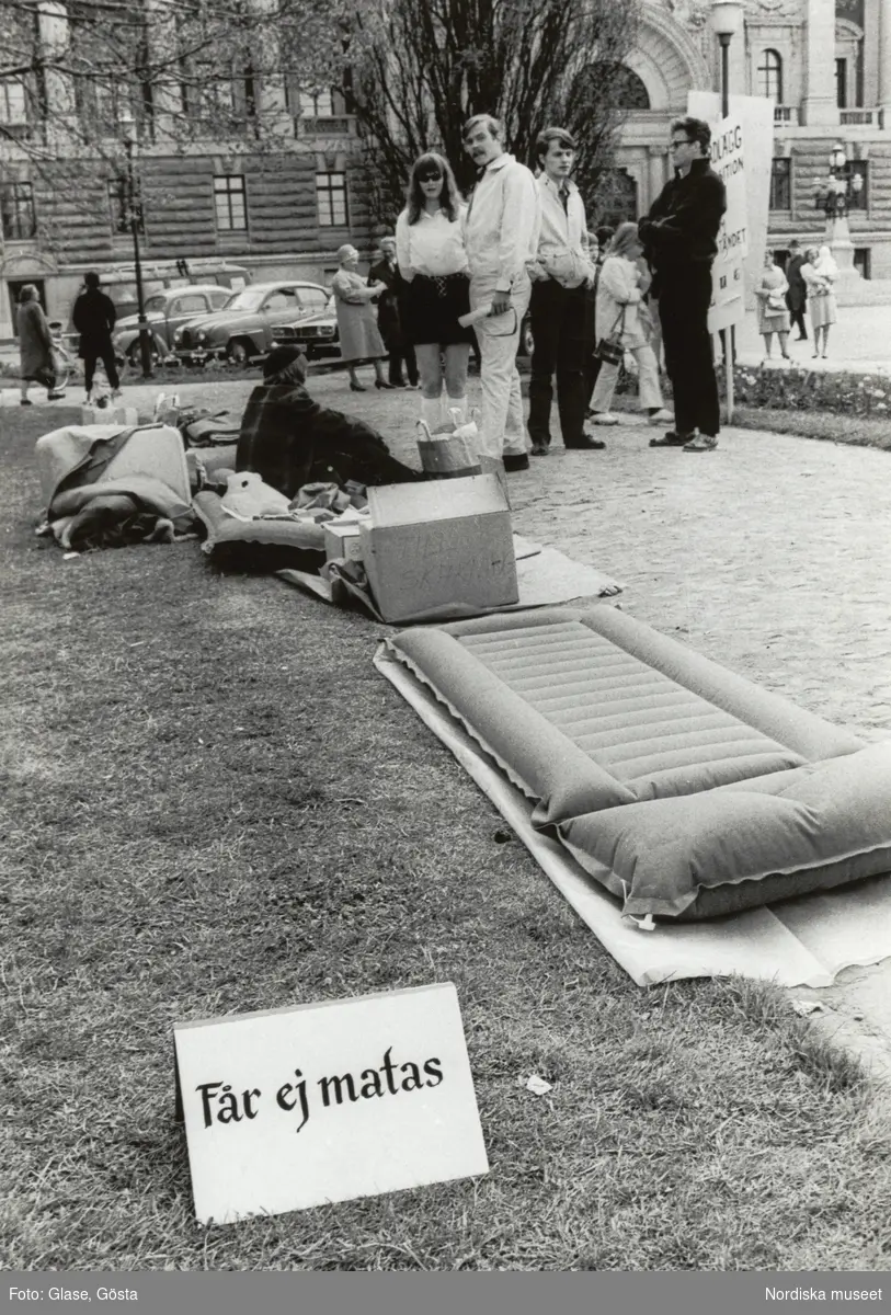 Kårhusockupationen vid Stockholms universitet i slutet av maj 1968. Utanför, på en gräsmatta har några personer slagit läger med luftmadrasser, filtar, kuddar, kartonger och kassar. En skylt med texten "Får ej matas" står intill på gräset. I bakgrunden syns gatumiljön med förbipasserande fotgängare och parkerade bilar.