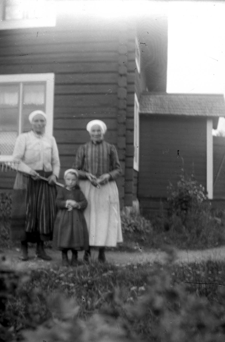 Gruppbild av två kvinnor och en flicka iframför ett knuttimrat hus. Den ena kvinnan håller en brödspade i handen.