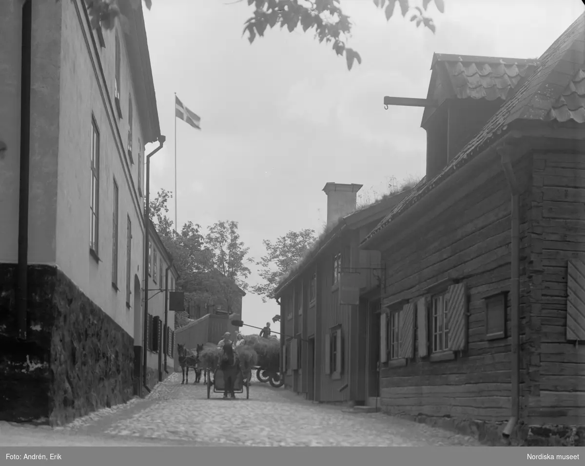 Gatubild från Skansen med Bokhantverkshuset till höger. På gatan hästdragna vagnar lastade med hö.