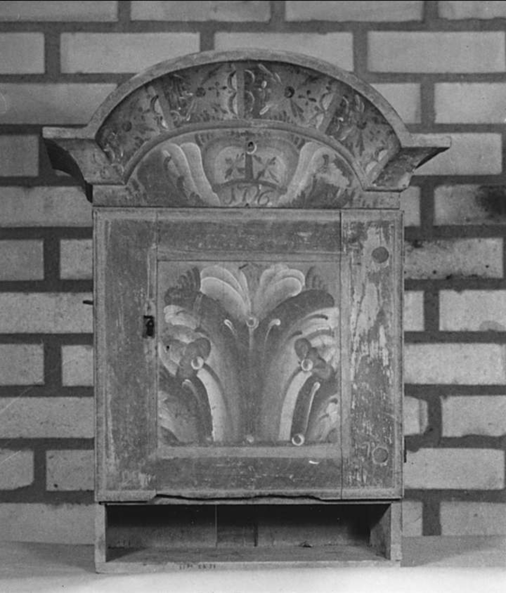 Hängskåp av trä. Brunmarmorerat med dekor i vitt, rött, svart och blått. Över dörren i kronan står: 1796. Inredning: en hylla och en skedhylla över den.