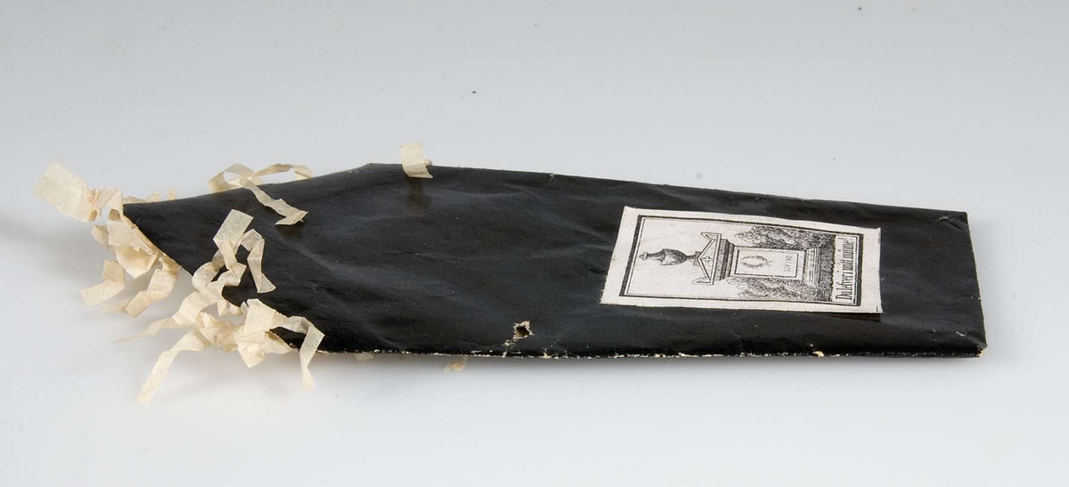 Begravningskaramell av svart glanspapper med vita fransar. Märke med gravvård och texten: "Du lefver i mitt minne".