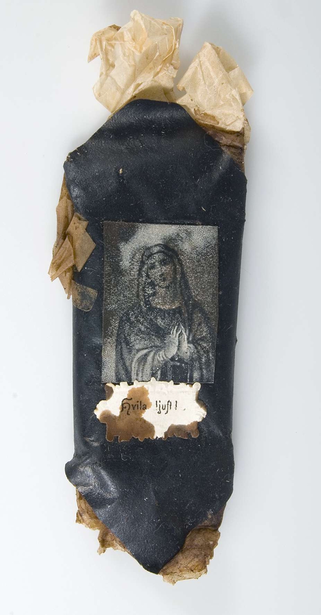 Begravningskaramell med omslag av beige silkespapper och svart glanspapper. Två pappersetiketter, den ena med tryckt bild av jungfru Maria, den andra med tryckt text: Hvila ljust!
