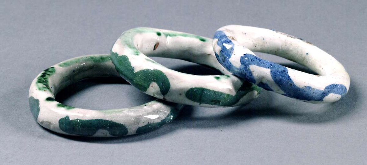 Tre gardinstångsringar av vitglaserat lergods. Två ringar med grönt mönster, en med blått.
