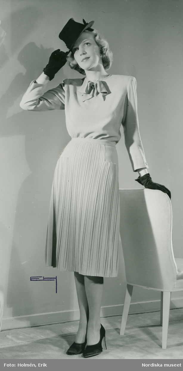 Helporträtt av kvinna i knytblus och kjol, samt hatt och handskar.