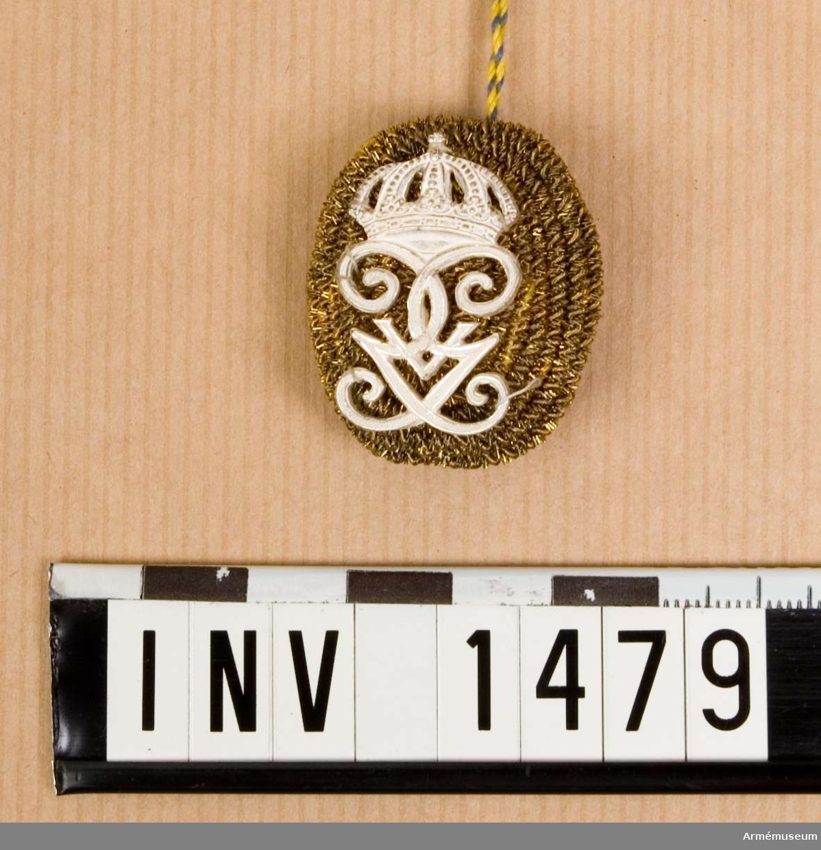 Oval, kupad med framsidan täckt av metallsnodd i guldfärg. G V:s namnchiffer i vitmetall påsytt. M/1908. På baksidan ett nät av guldtråd över metallstomme.  Hål för genomträdning av pompongnålen.