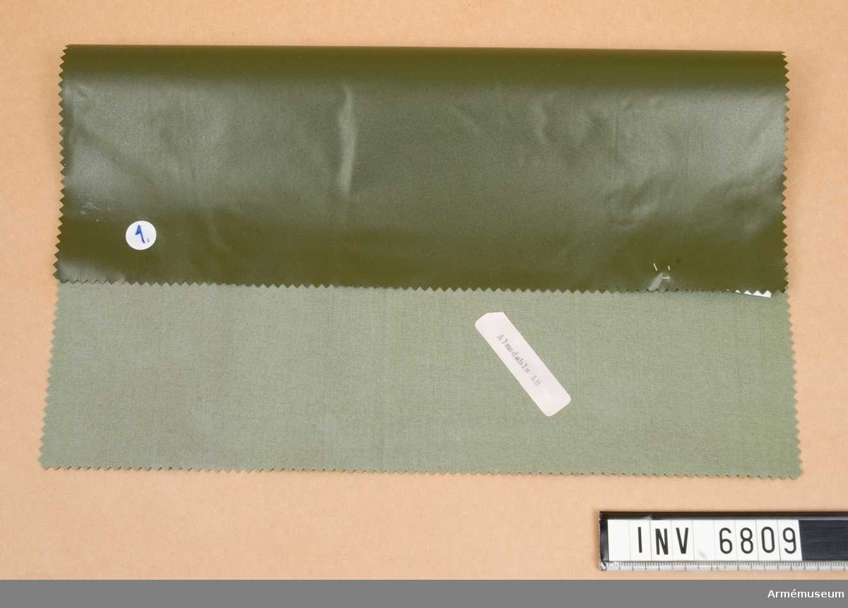 Prov av väv till regnkappa,  FMV. 1981. Mått 30O x 170 mm. Material avsedd för regnkappa. Nylon belagd med mjukgjord PVC i fastställd grön färg. Kvaliteten väger 240 gr per kvadratmeter.