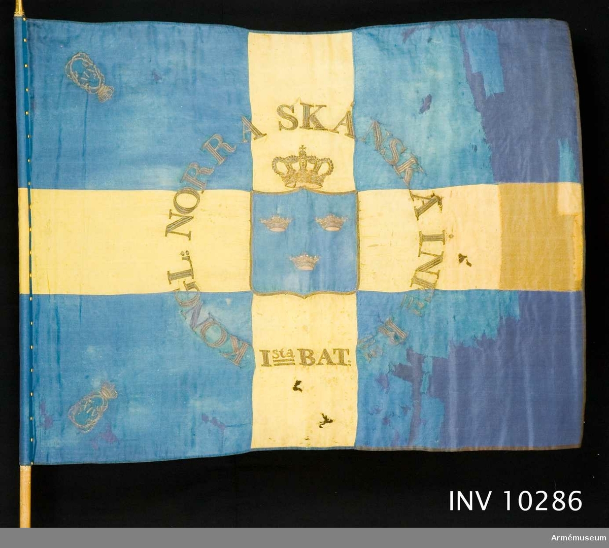Blå botten med gult kors. I guld broderat: "KONGL. NORRA SKÅNSKA INFri Rt Ista BAT." Stång av vitmålad furu, med spets. Spets har Carl XIV Johans namnchiffer.