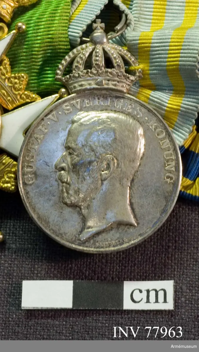 Grupp: M II.
Medaljen , omgiven av en upphöjd rand , har överst en stor kunglig krona, slutande i en kula, varigenom bandringen är trädd samt ett litet kors. Åtsidan: GUSTAF V SVERIGES KONUNG. Konungens huvud åt vänster, bart, UNDER AVSKÄRNINGEN: A LINDBERG. 
Frånsidan: Upptill: FEMTE OLYMPIADEN STOCKHOLM 1912 på 3 rader. Därunder en lagerkvist och en palmkvist lagda i kors samt Sveriges tre kronor inplacerade mellan kvistarna. Bandet; Ljusblått, vattrat med två smala gula ränder på varje sida. 