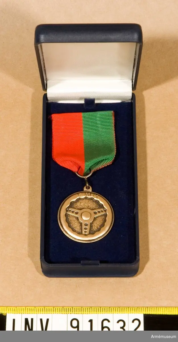 Etui innehållande Medalj.  FMKGM/SM.
På framsidan en ratt och text. Text även på baksidan.
Band till hälften grönt och till hälften rött.