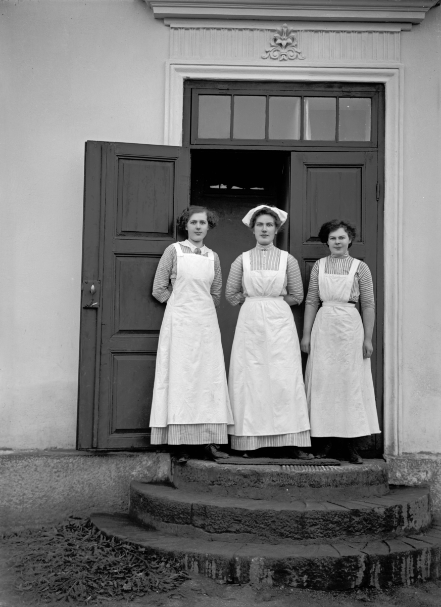 Tre kvinnor i arbetsklädsel framför entré (kökspersonal?)