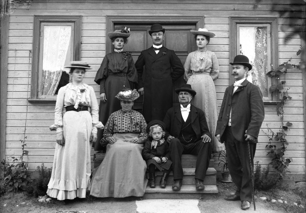 Fyra kvinnor, tre män och en flicka framför bostadshus, oidentifierad, troligen 13 augusti 1905