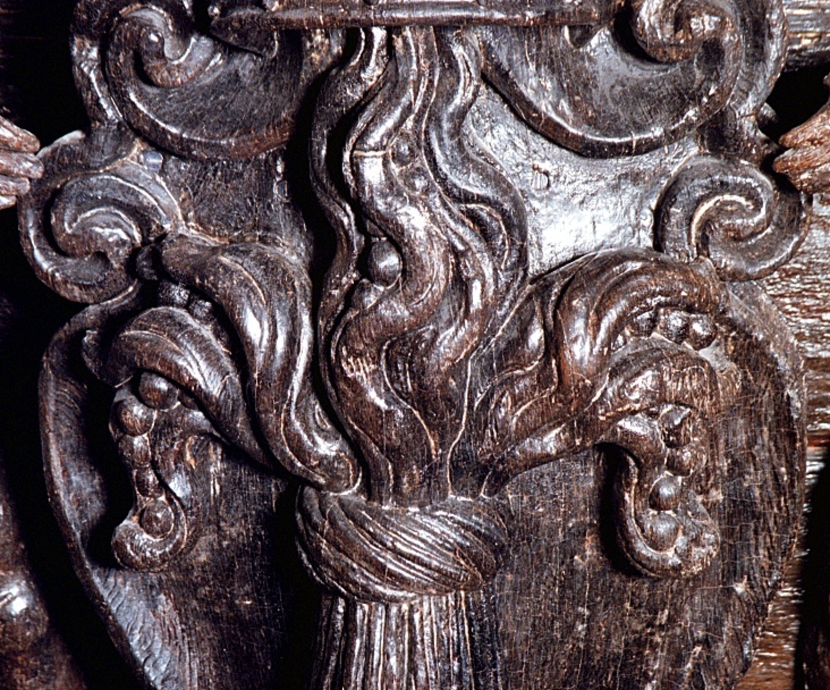 Skulpterad och profilerad sköld med kunglig krona och sädeskärve, vase. Skölden har förtjockade kanter och kraftiga rullverksformationer upptill och nedtill. Kärven är snidad med markerade strån och ax. Sköldens baksida är slät.

Text in English: Sculpted shield depicting the royal crown and a sheaf of corn, (called Vase in Swedish), carved in relief. The shield is carved with thickened edges and scrollwork formations at the bottom and top. The crown is shown in perspective and the stalks of the corn in the corn sheaf are clearly defined. The reverse side of the shield is smooth.