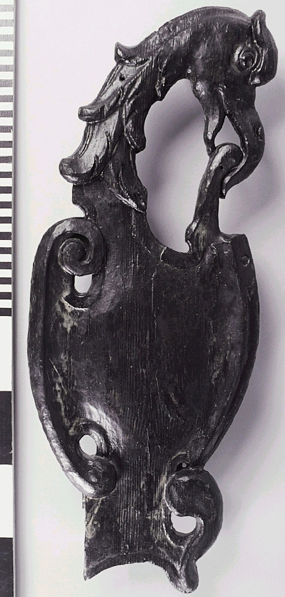 Skulpterad sköld med utsnidat fågelhuvud, med tre ursprungliga spikhål. Sköldens mittparti är konvext och har en oregelbunden och oval form, med asymmetrisk kantbård. Utmed den ena sidan avslutas bården upptill i en volut. Avsplittring på den nedre vänstra delen, annars väldigt välbevarad.

Övre delen av skölden är utdragen i ett stort och smalt, profilställt fågelhuvud med stiliserade halsfjädrar och stor, böjd näbb. Fågeln håller i sin näbb en rundad hörnflik från sköldens mittparti.

Det nedre partiet av skölden har ursprungligen sannolikt varit symmetrisk, med uppåtvridna, förtjockade flikar. Den ena sidan är dock bruten. Skölden, som delvis är genombruten, har en slät och jämn baksida. Mindre sprickor på sköldens mitt.

Text in English: Sculpted irregular shield decorated with a bird''s head.
The shield''s centre is convex and has a asymmetrical, oval shape, with an asymmetrical cord edging. On one side, the cord terminates upwards in a volute.

The top of the shield extends into a large narrow bird''s head in profile. It has stylized neck feathers and a large curved beak. In its beak, the bird is pulling and uncurling a scroll from the main body of the shield.

The lower ege of the shield probably terminated symmetrically when carved but one side is broken off. The lower edges are upturned and thickened. The shield is partly open-worked. The back is smooth and even.
The shield is in good condition, apart from the broken parts. A few minor cracks in the centre of the shield.