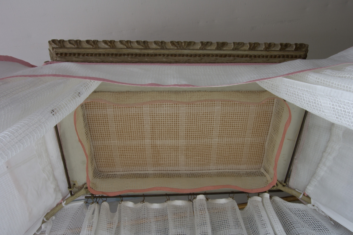 Sängklädsel i sju delar, tillverkad av handvävt tyg, bomullsgarn i varepn och lingarn i inslaget, vitt, vävt i stramalj med mönster i stora rutor, myggtjäll. Modell som är formsydd efter sängens överbyggnad, med nedfallande draperier på sängens baksida mot väggen. Sidstyckena upptill med foder. De är fastknutna med bomullsband i sängstommen. De främre förhängena är uppfästa med omtag. Under den lilla sänghimmeln hänger en äldre liten himmel av liknande tyg. På utsidan av himmeln sitter en kappa i tre delar över förhängena på sidor och fram. Fram- och bakhängena har metallringar i överkanten, som är trädda på metalltenar i sängstommen. Kapporna, omtagen och de sömmar som följer sängstommen är alla kantade med vävda band av rosa bomull.