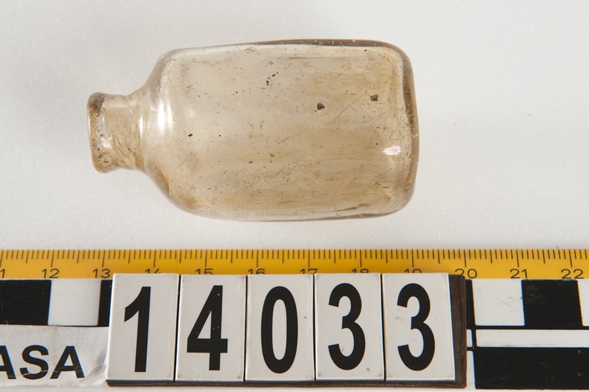 En gul-brun fyrsidig glasflaska.

Small glass medicine bottle. No damage to the artifact but the bottle has a misshapen shoulder from the manufacturing process. Glass is clear with a brownish tint, possibly potash-lime glass.
