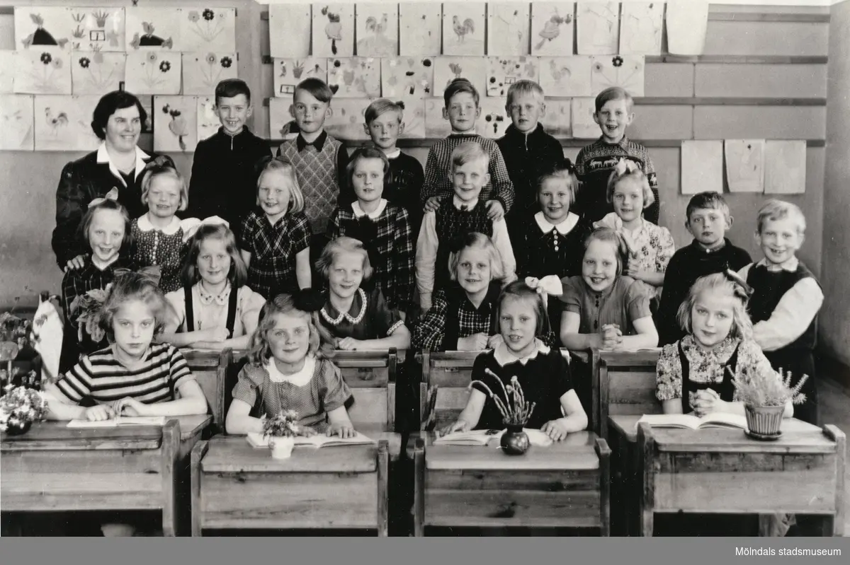 Fröken R. Sylvén med 1:a klass-elever på Centralskolan 1943-44. Flickor sittandes i bänkar. Flickor och pojkar ståendes bakom. Teckningar på väggen. Med på bild finns, sittandes till höger, Berit Gustafsson (född Larsson 1936).