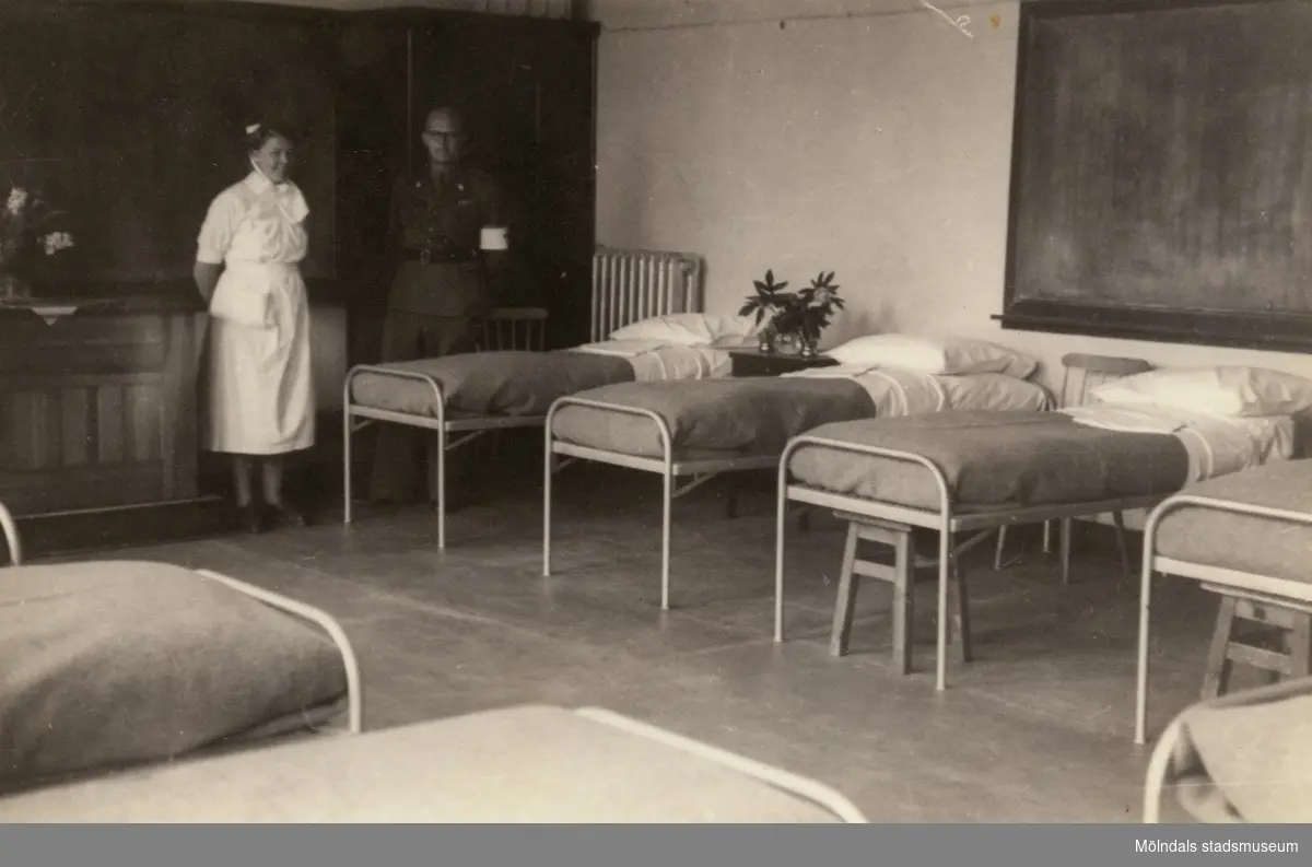 "Karantänsförläggning" på Kvarnbyskolan för överlevande från koncentrationsläger i Tyskland och Polen 1945.  
Skolan fungerade under denna sommar som beredsskapssjukhus.
 
En sjuksal med sängar samt en sköterska och en man från Röda Korset.
Den svarta griffeltavlan bakom sängarna till höger visar på att det är en av skolsalarna som gjorts om till sjukhussal.