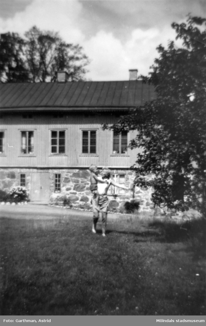 Astrid Garthmans syster Ingrid Karlsson håller sin son Lars i famnen. Man ser Solbacken eller "Skogsvaktarebostaden" i bakgrunden. Gunnebo, 1960-tal.