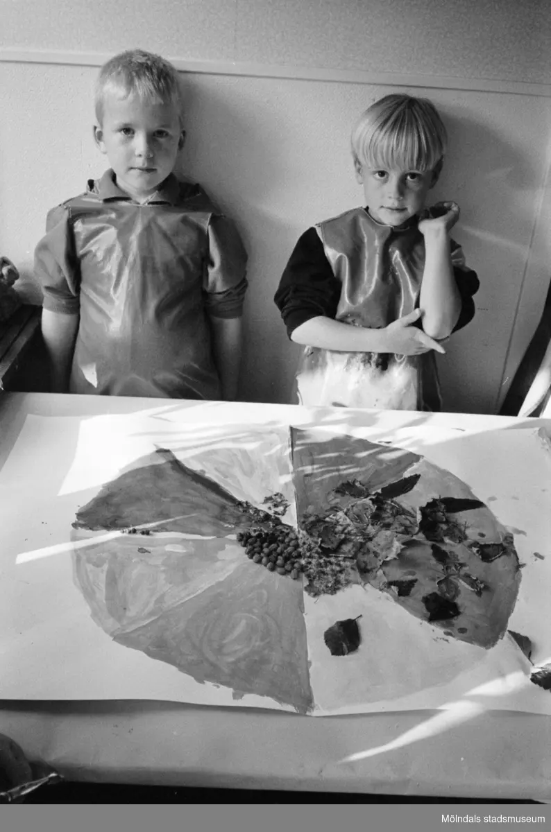 Två pojkar, iklädda förkläde, står uppställda bakom ett bord där det ligger en stor målad teckning, dekorerad med bl.a blad. Båda gossarna tittar mot kameran. Katrinebergs daghem, 1992-93.