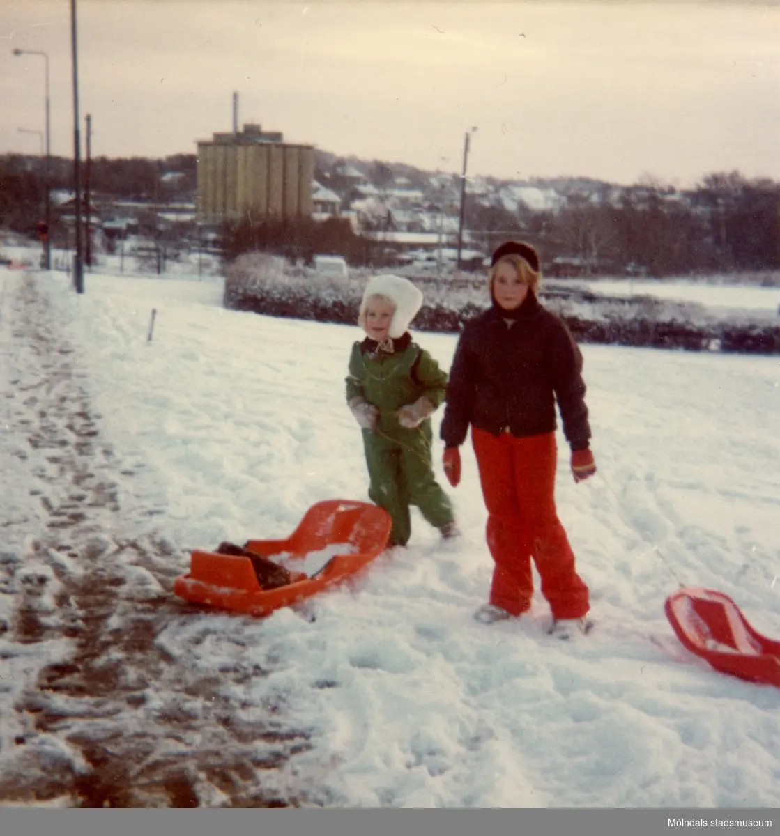 Christine och Ingela med pulkor vid Delbancogatan i Mölndal, december 1976.
Den gamla SOAB-cisternen i bakgrunden.