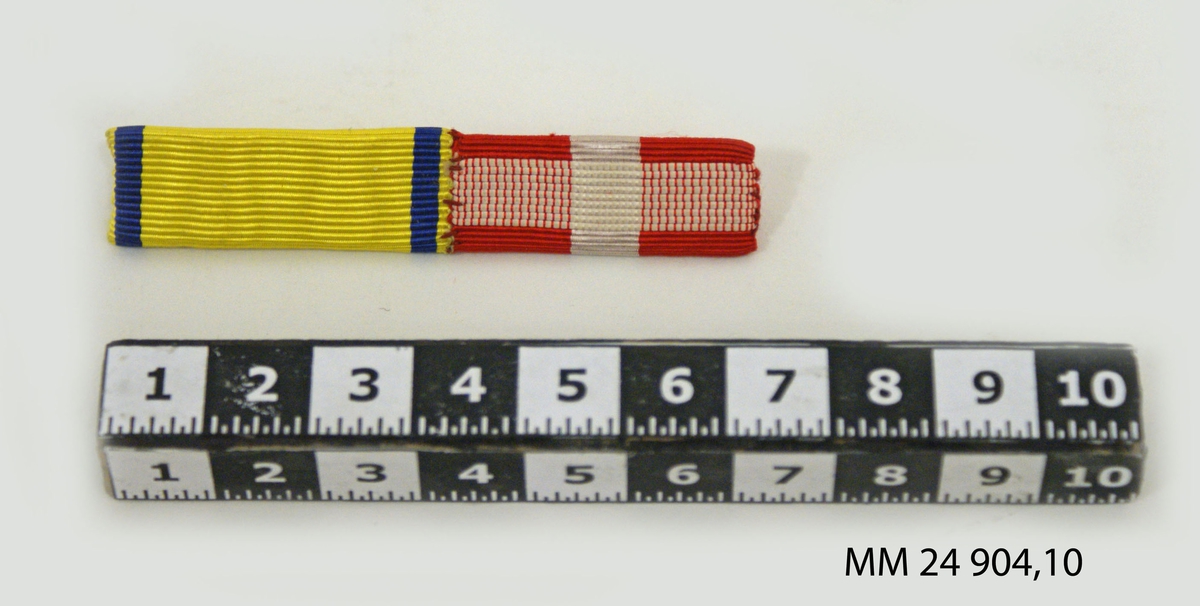 Släpspänne av räfflat sidenband på rektangulär bricka. En del i lodrät randning blått, gult, blått, som står för kungliga svärdsorden. En del i färgerna rött och vitt, som är en dansk medalj (möjligen för räddning av drunknande). Fästnål på baksidan.