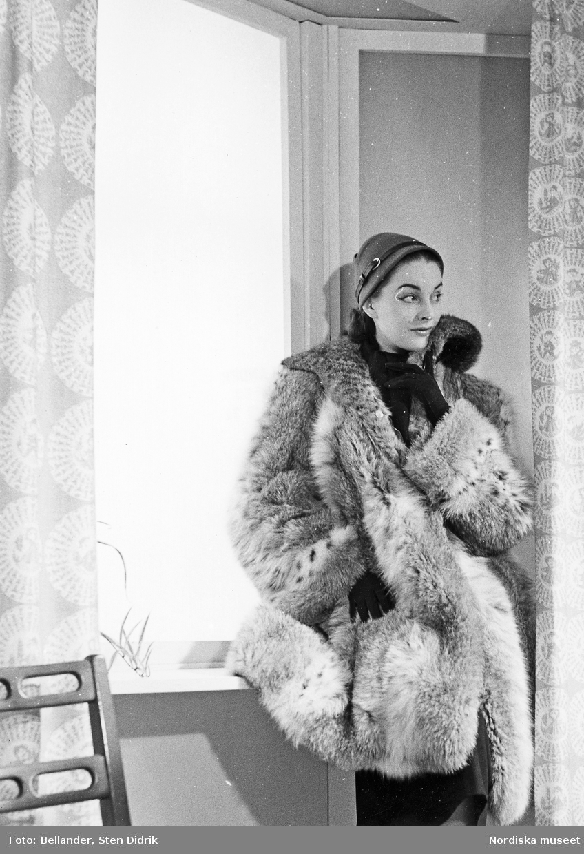Modell, möjligen Anna-Lisa Olsson,  i pälsjacka, hatt och handskar, vid ett fönster.