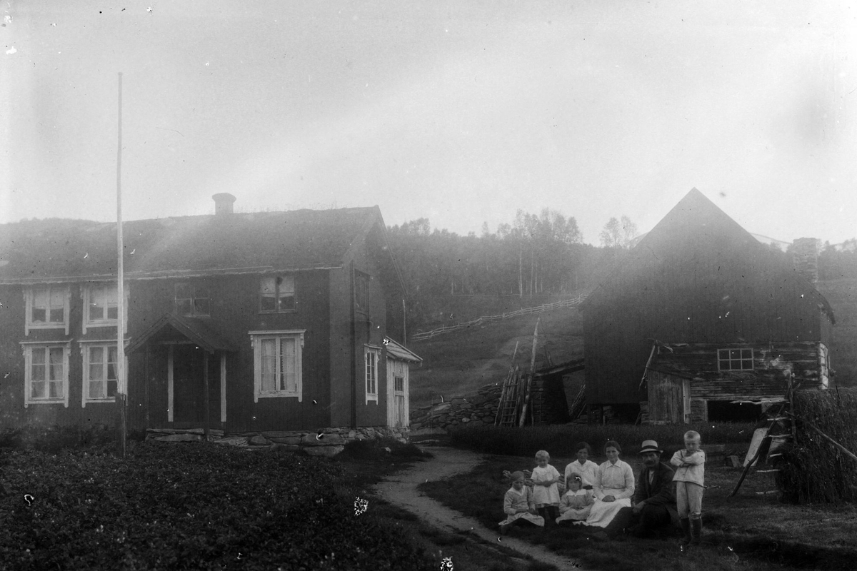 Gårdstun med hus, fjøs og et steingjerde. Til høyre er en gruppe personer, trolig familien som bor på gården.