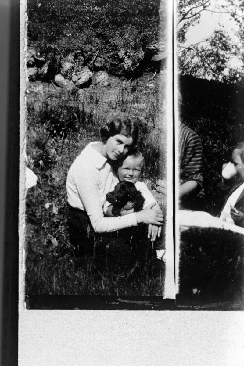 Reproduksjon av bilde tatt utendørs, med en kvinne med en gutt og hund på fanget. Til høyre sees kanten på et annet fotografi.
