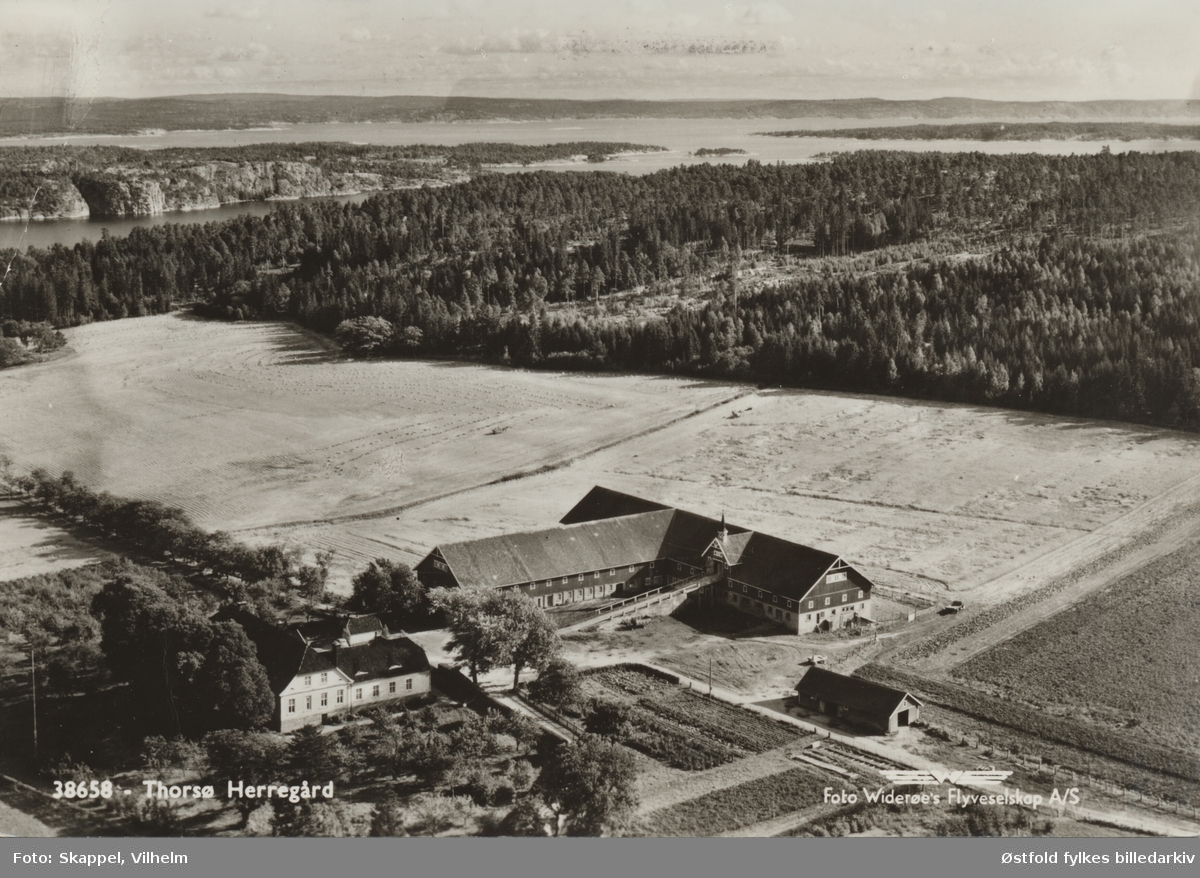 Thorsø Herregård i Borge, med uthus og åkre. I bakgrunnen ses fjorden. Postkort.