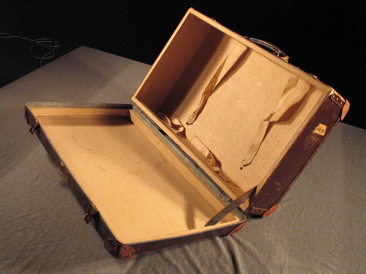 Låsbar pappkoffert med papirtrekk inni. Skinnlappar som forsterkningar på hjørna. Handtak i metall.