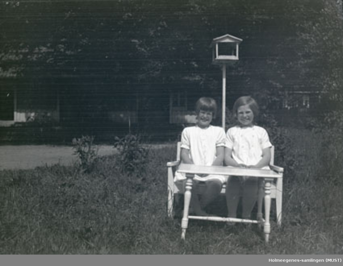 To jenter på en liten benk i hagen på Holmeegenes i Stavanger. Det står et fuglebrett bak dem, og huset skimtes i bakgrunnen.