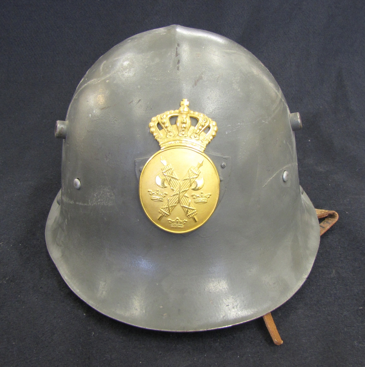 Stålhjälm för polisman (modell 1941). Polisemblem med krona i gulmetall. På emblemet, korslagda fasciner och tre kronor.