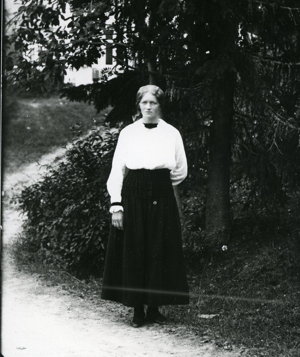 Kvinne i helfigur, kledd i hvit skjorte og mørkt skjørt, fotografert på vei med skog og hvit bygning i bakgrunnen