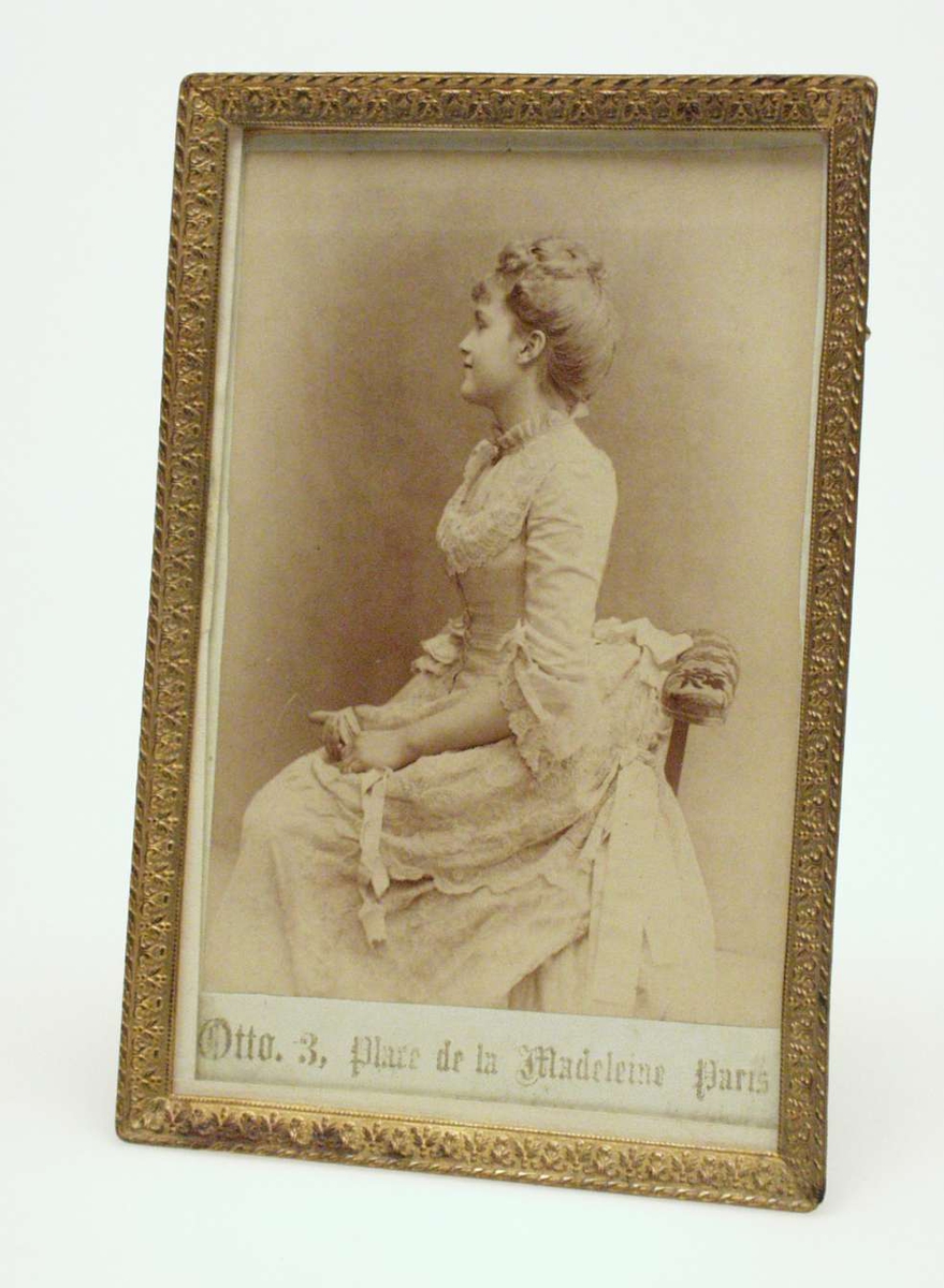 Ung kvinne med oppsatt hår og hvit kjole i damask og kniplinger. Stolen har polstret armlene. Bildet er tatt i profil.