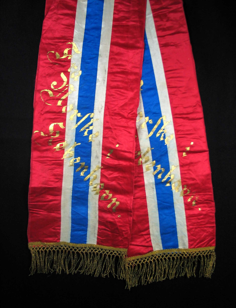 Begravelsessløyfe i rød, hvit og blå silke med frynser og tekst i gull.
