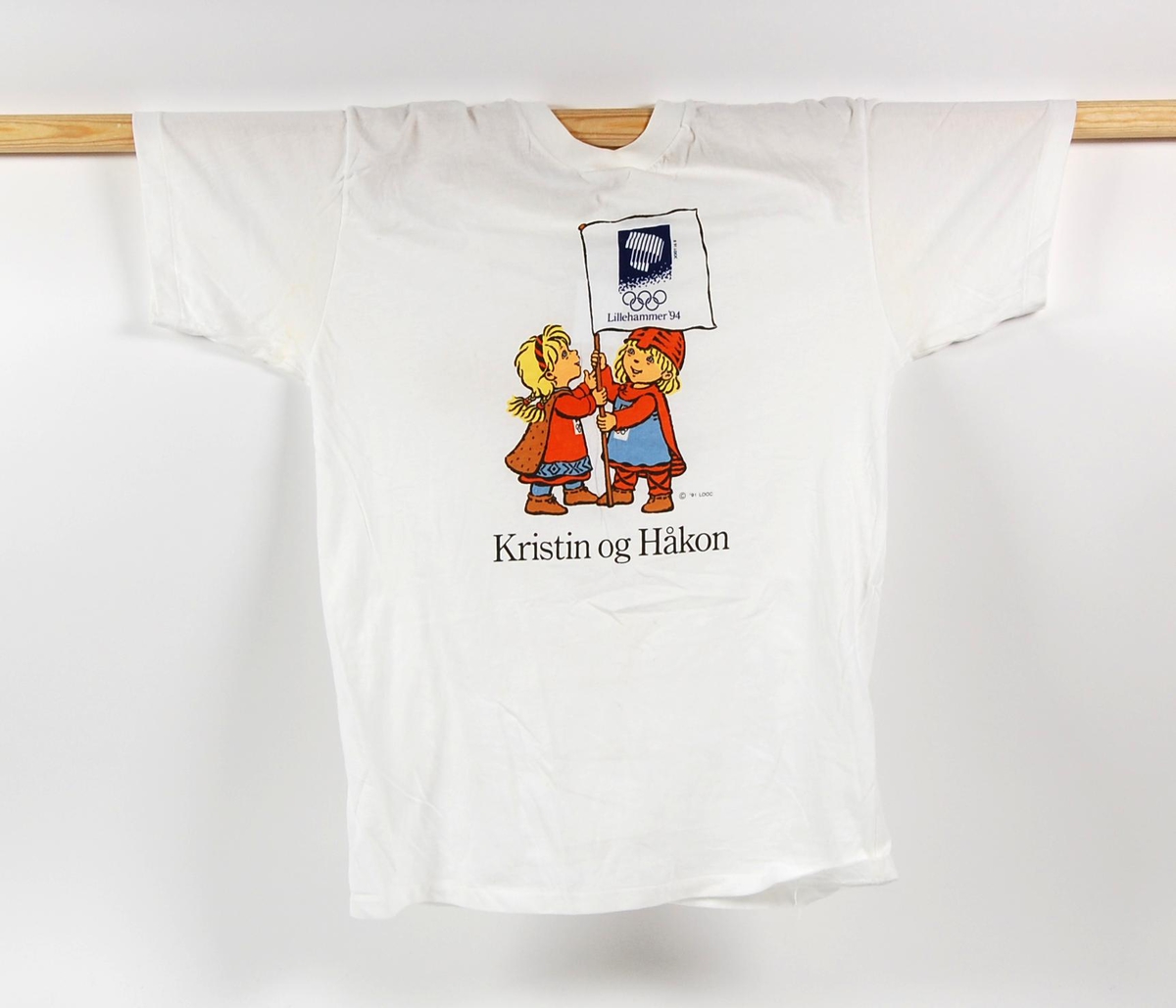 Hvit t-skjorte med flerfarget motiv av maskotene for de olympiske vinterleker på Lillehammer i 1994, Kristin og Håkon. Kristin og Håkon holder et flagg med logo fro de olympiske vinterleker på Lillehammer i 1994. T-skjorten er i størrelse L.